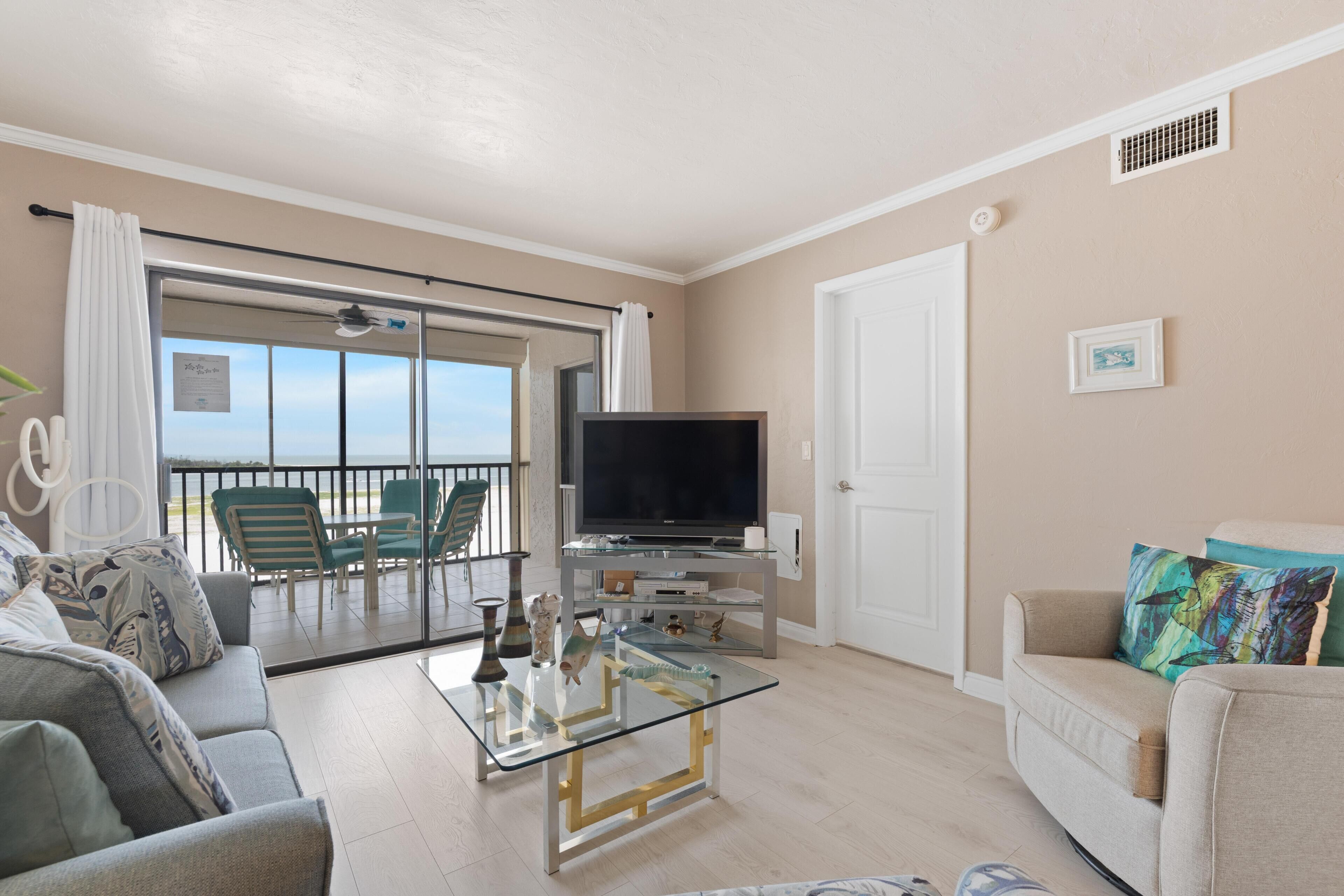 Ein Wohnzimmer mit Glastisch, einer Couch, einem Fernseher und einem Balkon mit Sitzgelegenheiten.