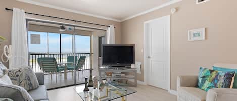 A living room with a glass coffee table, a sofa, a television, and a balcony with chairs.