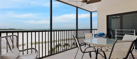 A balcony with a view of the beach, a table with chairs, and a ceiling fan.