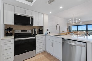 A modern kitchen with stainless steel appliances, white cabinetry, and a large window offering a view. - Carlos Pointe #231 Beach Bum Getaway (Fort Myers Beach)
