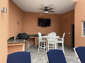 A patio with a ceiling fan, a TV mounted on the wall, a grill, and a table with chairs. - Cay Syrah! 4 bedroom home! Solar/battery backup! (Cruz Bay)