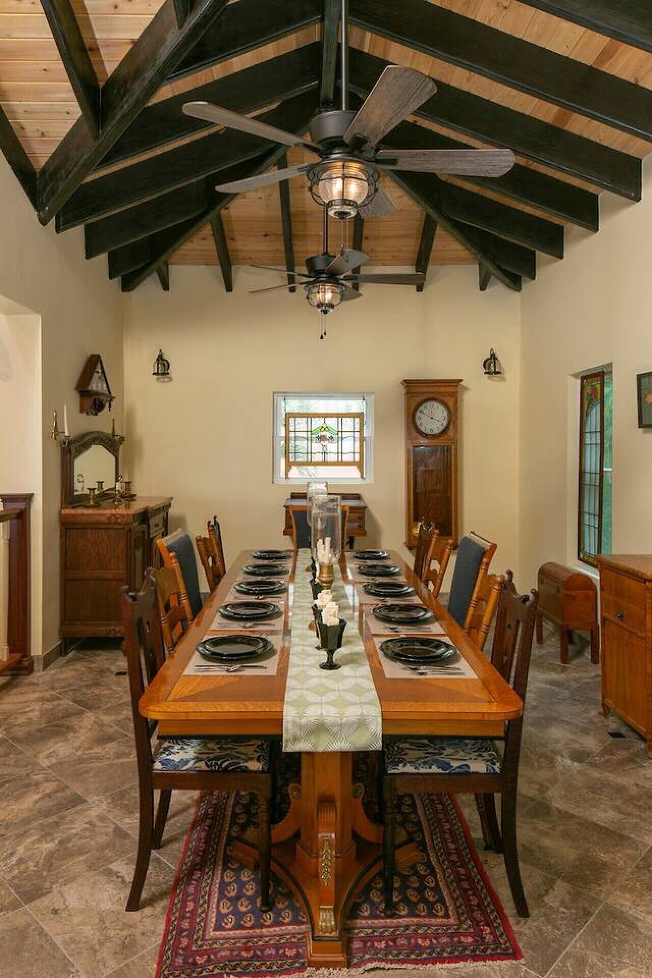 A dining room with a long wooden table, chairs, a chandelier, a clock, and a window.