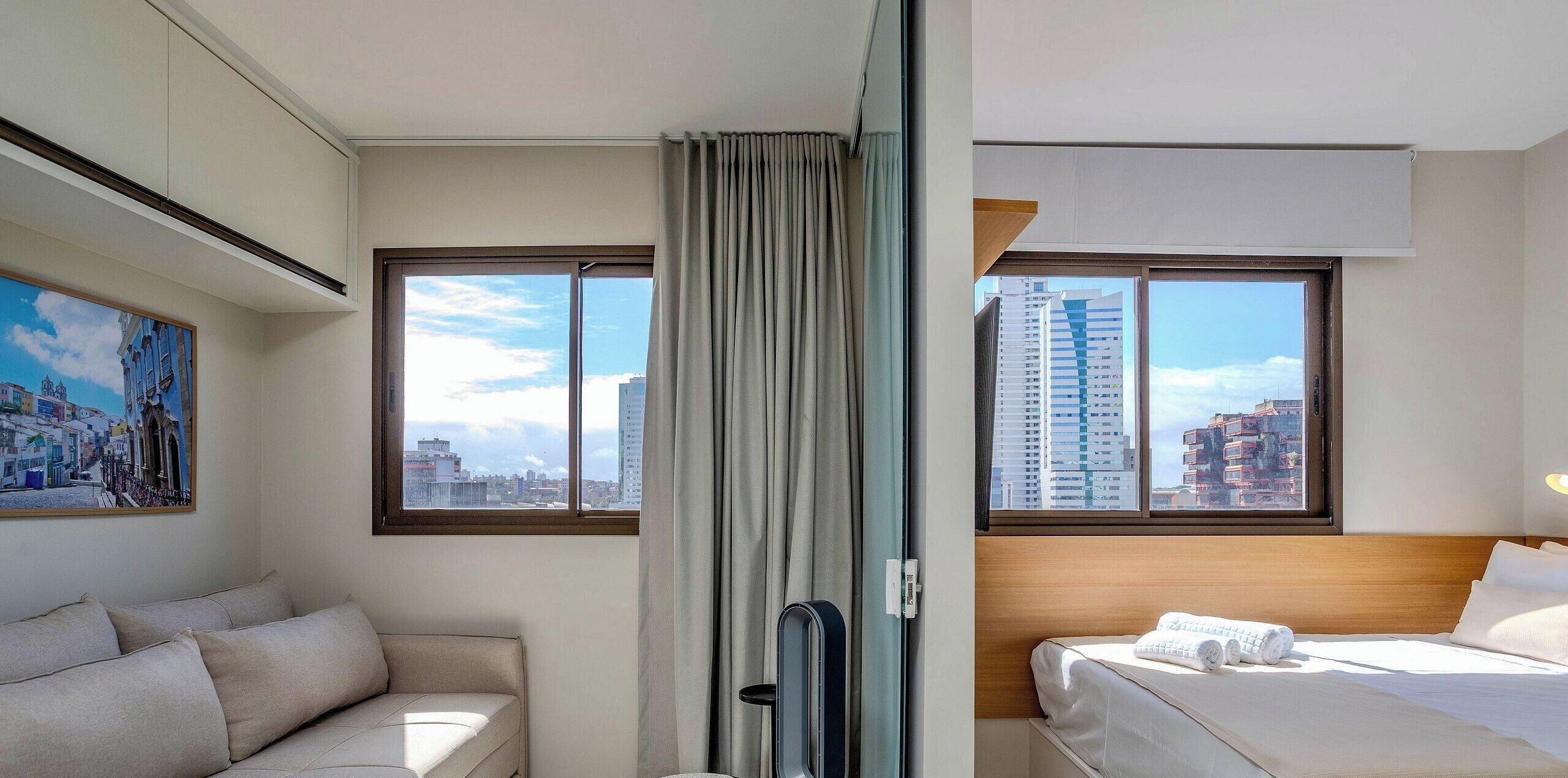 Ett hotellrum med en säng, en soffa, ett stort fönster med utsikt över staden och en tavla på väggen.
