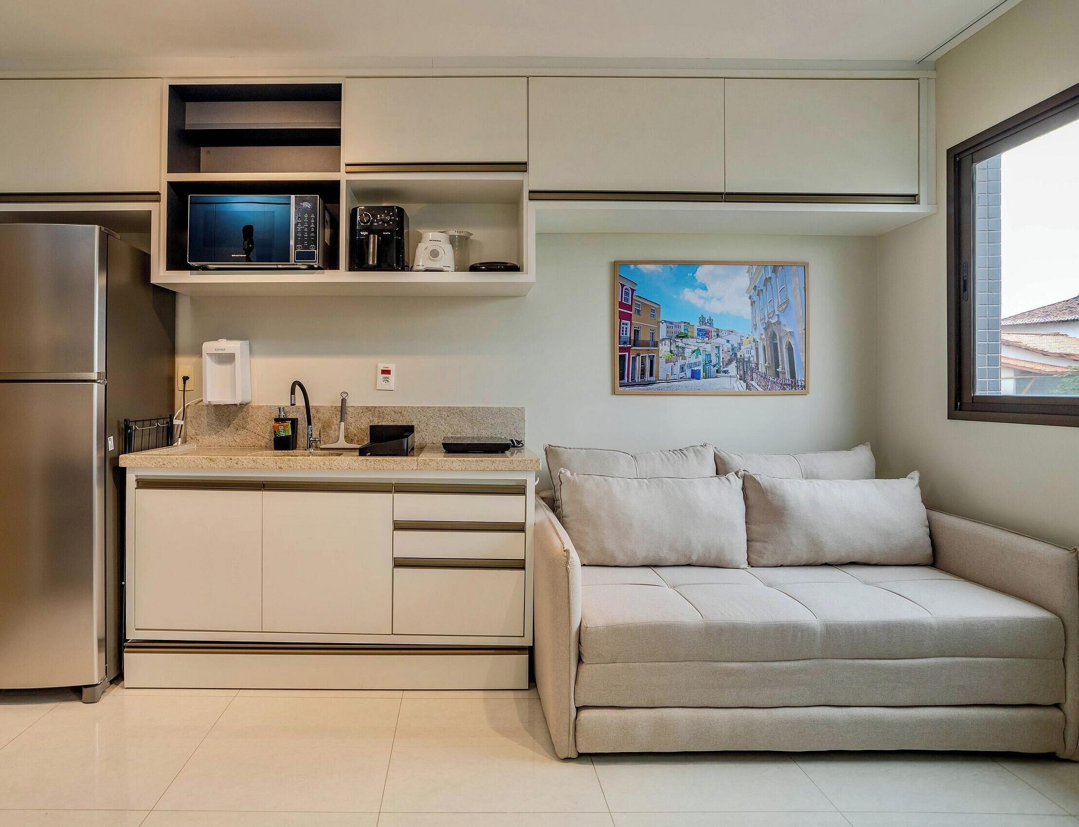 A compact kitchen with a built-in microwave, a coffee machine, and a toaster. There is a sofa with cushions and a painting of a cityscape on the wall.