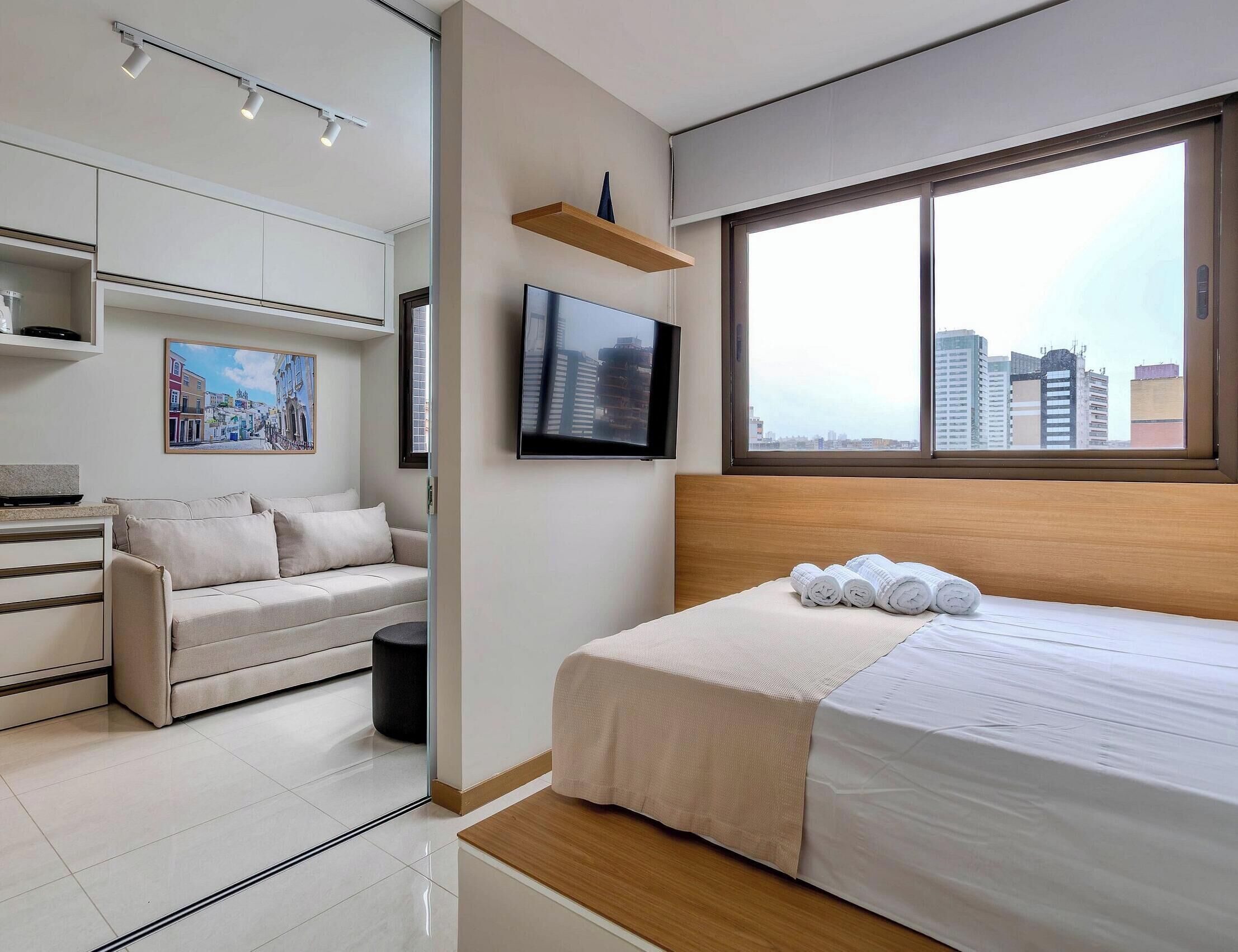 A modern hotel room with a bed, a sofa, a TV, and a city view.