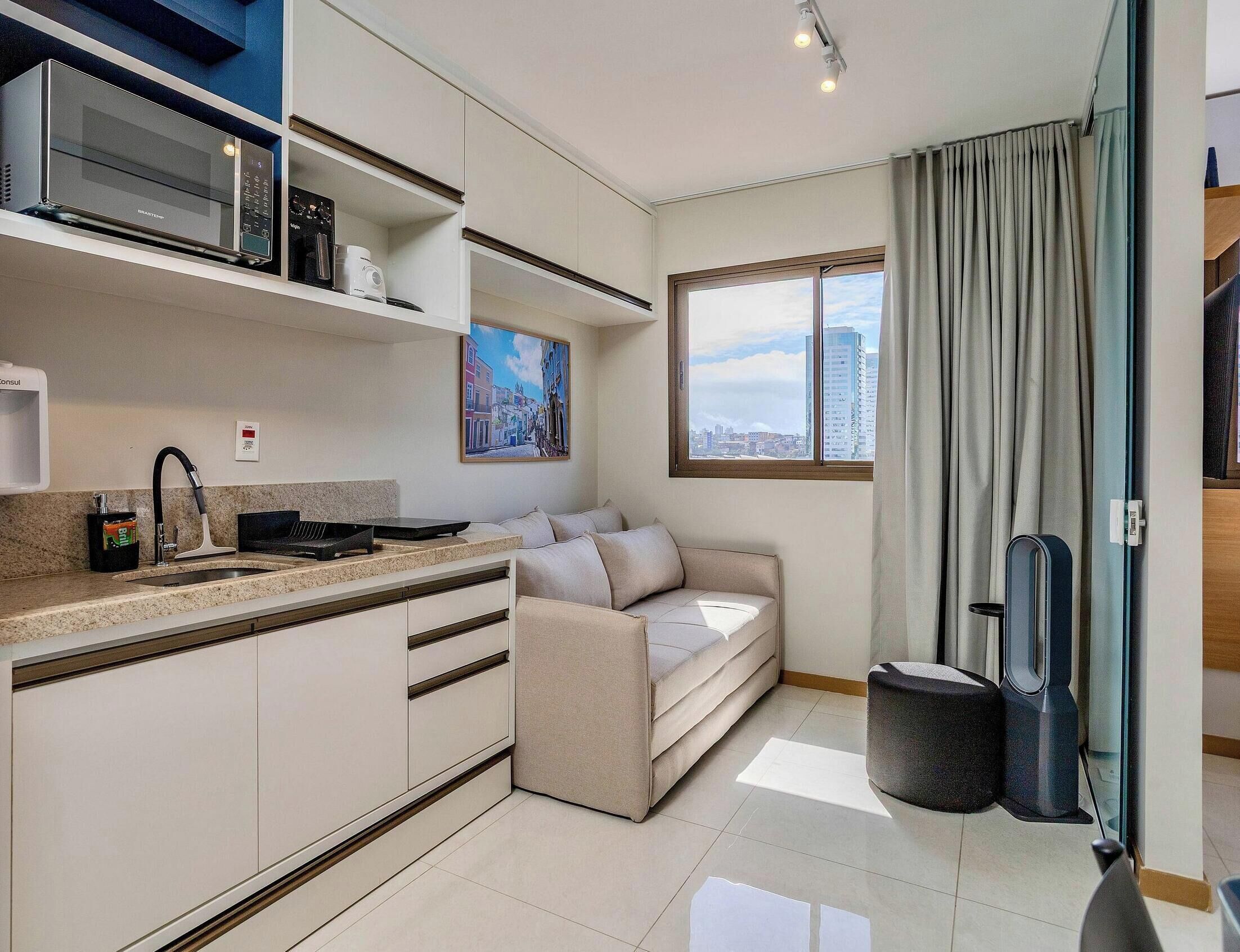 A modern kitchen with a built-in microwave, a coffee machine, and a sink. There is a sofa and a window with a city view.