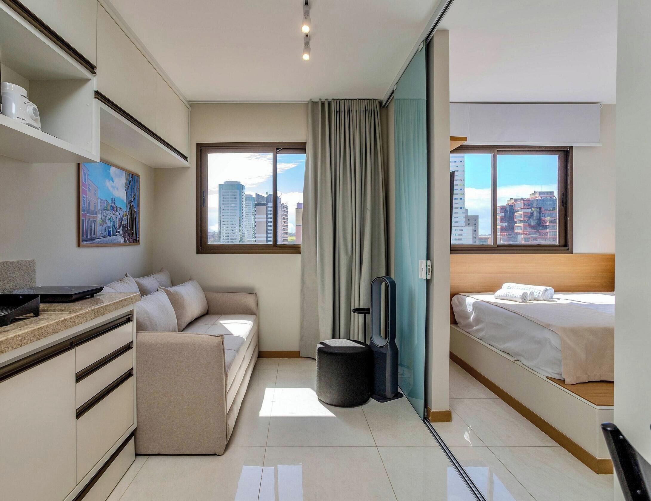 A hotel room with a bed, a sofa, a desk with a laptop, a window with curtains, and a city view.