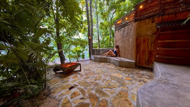 A stone patio with a fire feature, surrounded by lush greenery and a wooden structure.