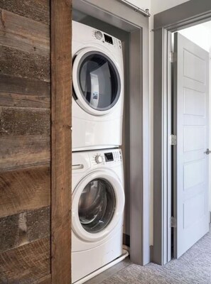 A stacked washer and dryer unit in a hallway with a wooden accent wall and a white door. - Bozeman Home with Private Fishing Access (Bozeman)