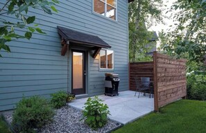 A house with a dark blue exterior, a front door, a small patio with a grill, and a wooden fence. - Bozeman Home with Private Fishing Access (Bozeman)