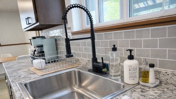 A kitchen with a stainless steel sink, a dish rack, a coffee maker, and a window with a view of trees. - Family-friendly duplex near Fargodome, kings+bunk (Fargo)