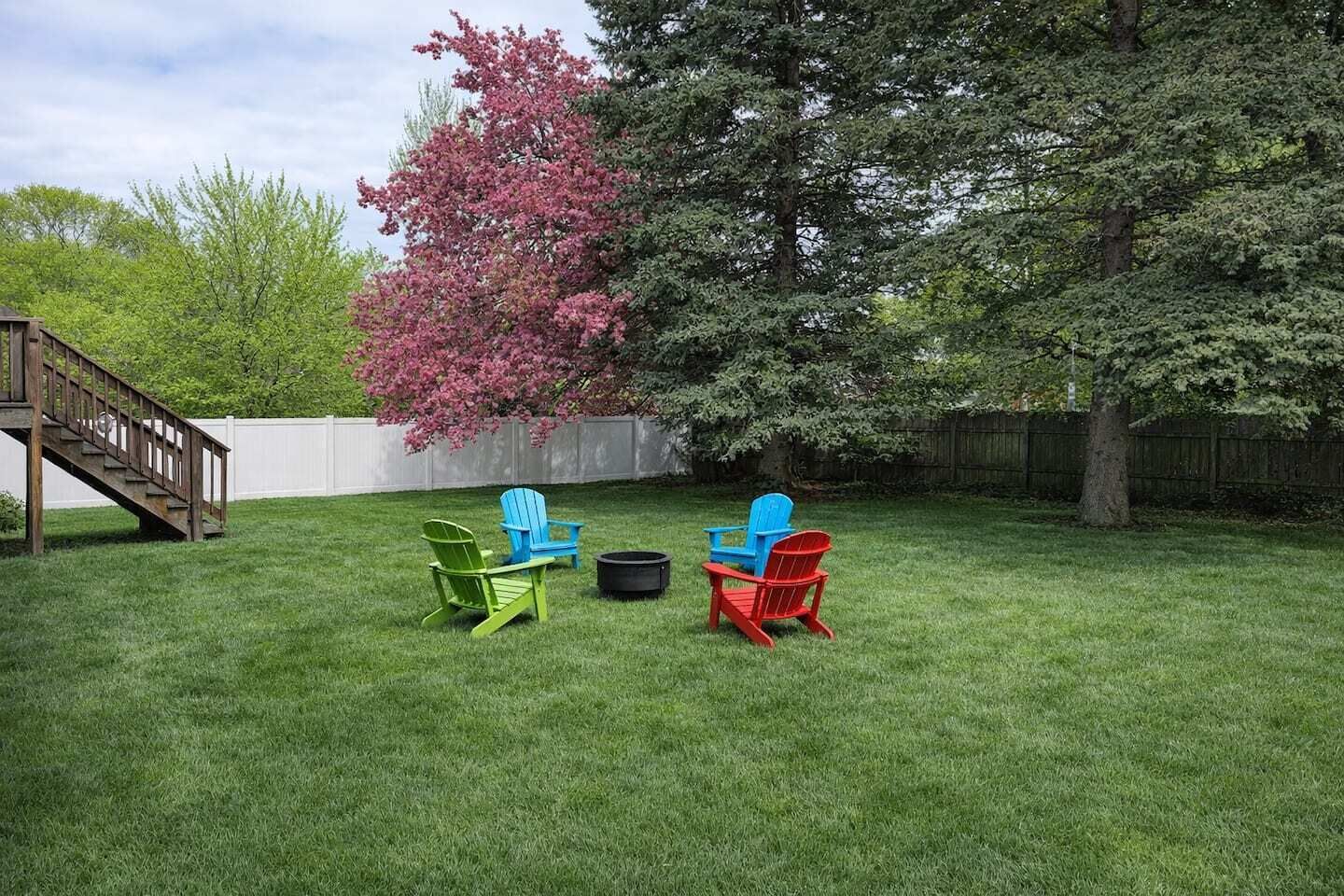 Un patio trasero con una terraza de madera, un árbol con flores rosadas y tres sillas Adirondack.