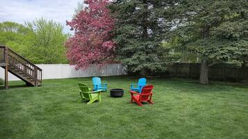Un patio trasero con una terraza de madera, un árbol con flores rosadas y tres sillas Adirondack.