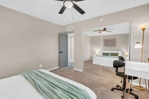 A bedroom with a bed, a desk, a chair, a ceiling fan, a mirror, and a door leading to another room. - Midtown Cozy Getaway! (Tucson)