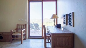 A room with a wooden desk, two chairs, a lamp, and a sliding glass door leading to a patio with chairs. - Exquisite Starr Pass Desert Haven with Pool (Tucson)