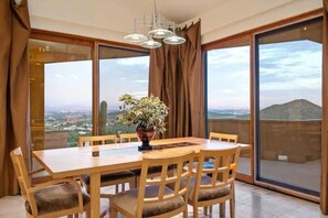 A dining area with a wooden table and chairs, a potted plant, and large windows offering a view of a landscape. - Exquisite Starr Pass Desert Haven with Pool (Tucson)