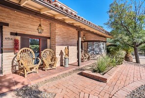 A building with a brick facade, numbered 6914, featuring outdoor wicker chairs, a covered patio, and a landscaped garden with cacti. - Southwest Gem-Pool & Mountain Views (Tucson)