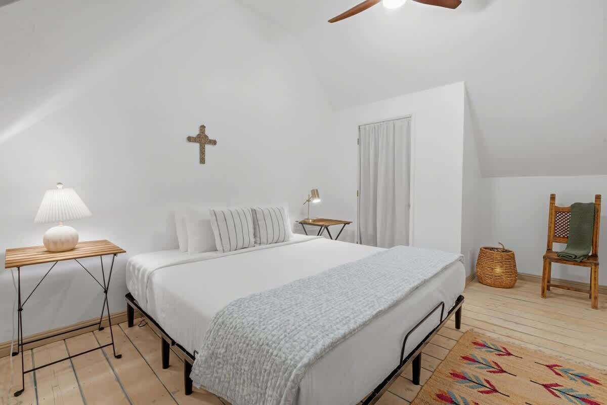 A bedroom with a bed, bedside tables, a cross on the wall, a window with curtains, a ceiling fan, and a chair.