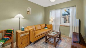 A living room with a sofa, a wooden side table, a coffee table, a lamp, a TV, and a window with curtains. - Walkable Barrio Charm (Tucson)