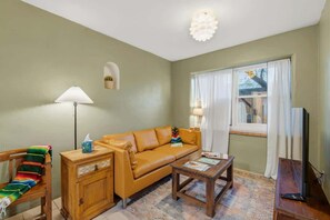 Ein Wohnzimmer mit einer Couch, einem Holztisch, einem Couchtisch, einer Lampe, einem Fernseher und einem Fenster mit Vorhängen.