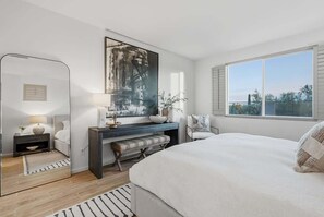Een slaapkamer met een bed, nachtkastje, lamp, kunstwerk, bankje en een groot raam.