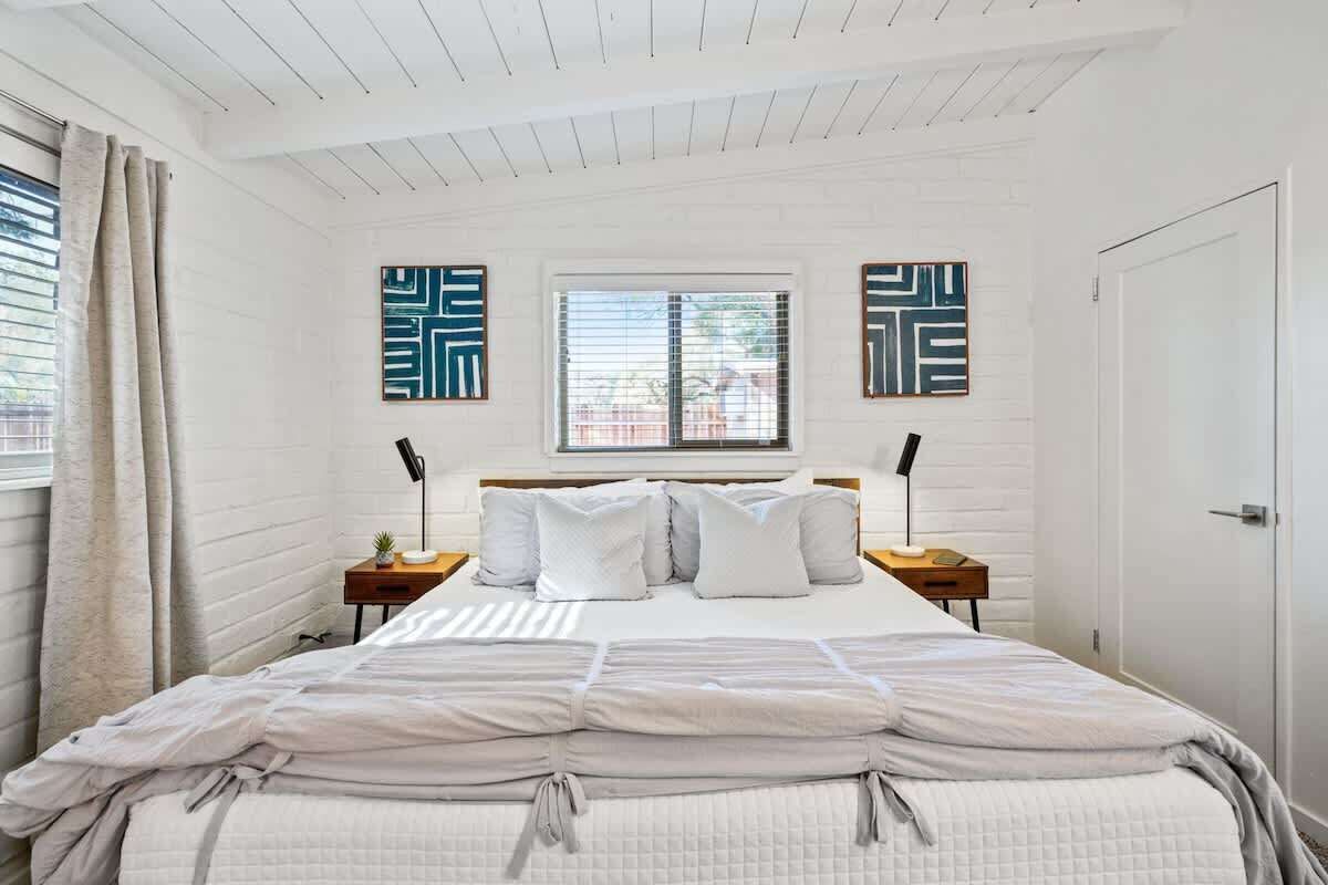 A bedroom with a bed, two bedside tables, a window with blinds, and a door.