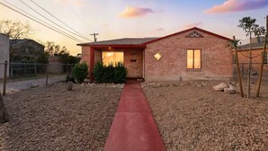 Una casa de un piso de ladrillo con un sendero rojo que lleva a la entrada, rodeada por un patio seco y pedregoso.