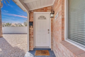 A white door with a number plate, a doormat, and a brick wall. - Sam Hughes Escape-Walk to U of A (Tucson)
