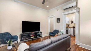 Ein Wohnzimmer mit Flachbildfernseher, einem grauen Sofa, einem blauen Sessel, einem kleinen Tisch mit Pflanze und einem Bücherregal.