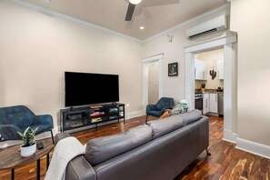Ein Wohnzimmer mit Flachbildfernseher, einem grauen Sofa, einem blauen Sessel, einem kleinen Tisch mit Pflanze und einem Bücherregal.