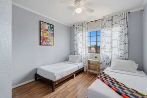 A room with two single beds, a ceiling fan, a window with curtains, a small table with a plant, and a colorful painting on the wall. - Bright & Spacious Family Retreat (Tucson)