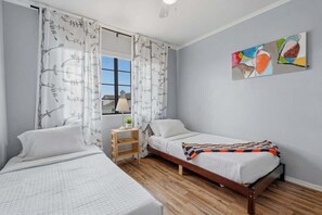 Ein Schlafzimmer mit zwei Betten, einem Fenster mit Vorhängen, einem Nachttisch mit Lampe und einem an der Wand hängenden abstrakten Gemälde.