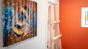 A room with a painting of a bee on the wall, a wooden ladder leaning against a white door, and an orange wall. - Bright Desert Retreat Pet Friendly (Tucson)