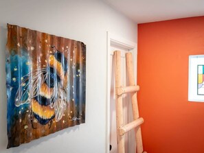 Ein Zimmer mit einem Bienenbild an der Wand, einer an einer weißen Tür angelehnten Holzleiter und einer orangefarbenen Wand.