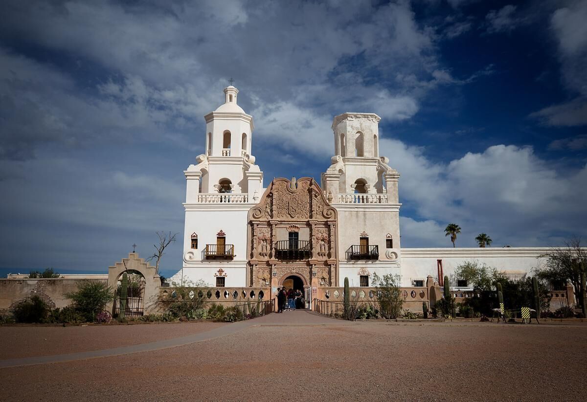 A large white building with two towers and a central entrance, surrounded by a paved area and cacti.