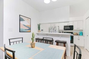 Une cuisine moderne avec des meubles blancs, un four à micro-ondes et un évier. On y trouve une table à manger, une plante et un tableau accroché au mur.
