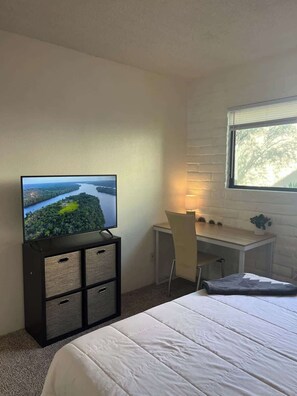 Une chambre à coucher avec un lit, un bureau avec une chaise, un téléviseur posé sur un meuble et une fenêtre donnant sur un paysage agréable.
