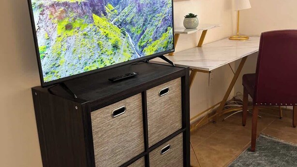 Et fjernsyn på et skab, et skrivebord med en lampe, og en stol i et rum.