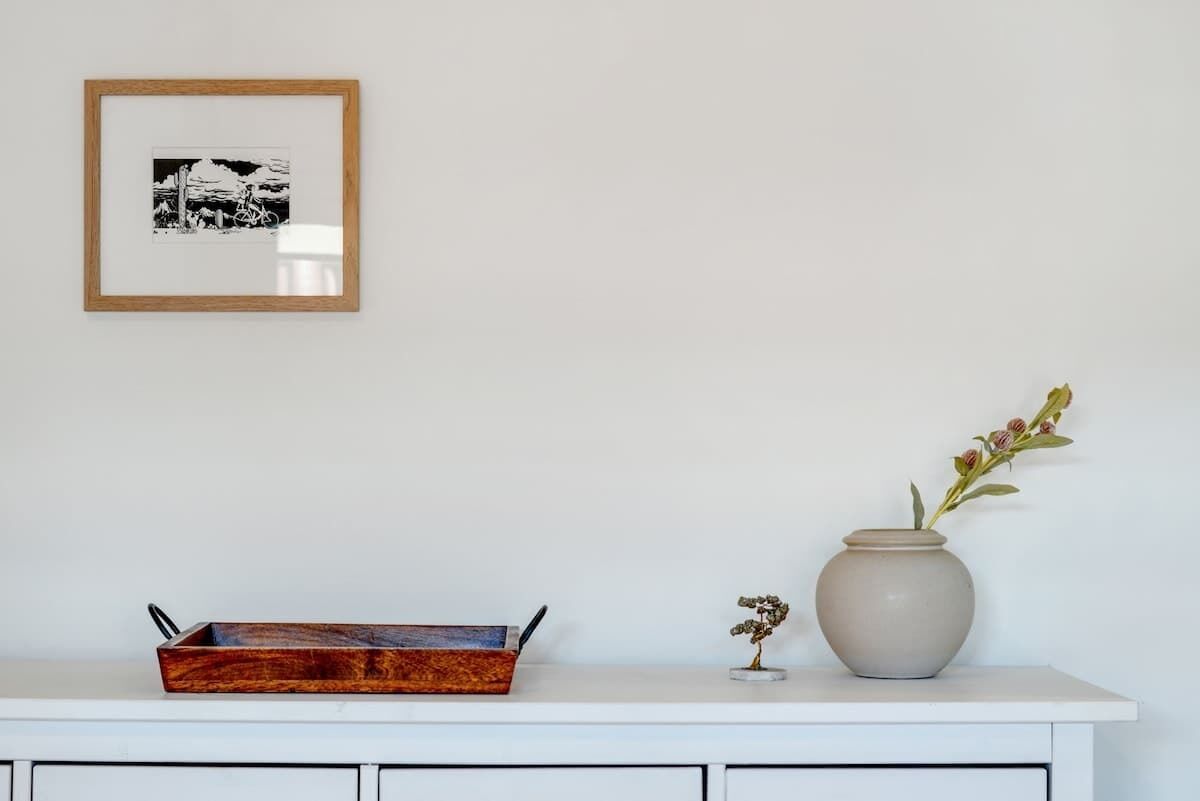 A white cabinet with a framed picture, a wooden tray, and a vase with flowers.