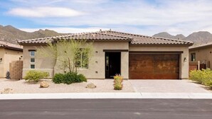 A modern two-story house with a garage, surrounded by a landscaped garden with rocks and shrubs. - Gated Desert Oasis w/ Pool (Marana)
