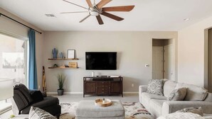 A living room with a ceiling fan, a flat-screen TV, a sofa, a coffee table, and a potted plant. - Gated Desert Oasis w/ Pool (Marana)