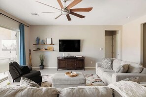 A living room with a ceiling fan, a flat-screen TV, a sofa, a coffee table, and a potted plant. - Gated Desert Oasis w/ Pool (Marana)