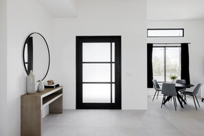 Ein modernes Zimmer mit einem großen Fenster mit schwarzem Rahmen, einem runden Spiegel, einem Holzschreibtisch und einem Essbereich mit Stühlen und Tisch.
