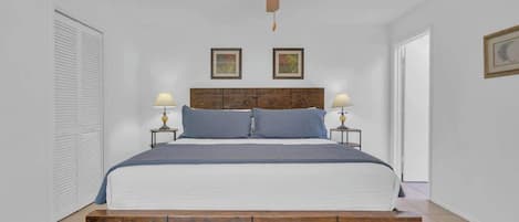 Un dormitorio con cabecera de madera, dos mesitas de noche con lámparas, un ventilador de techo y una puerta blanca.