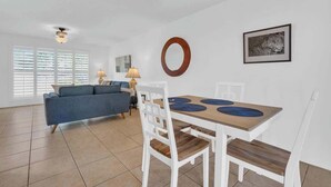 A modern living room with a sofa, dining table, and chairs. There are windows with blinds, a ceiling fan, and a framed picture on the wall. - Warm & Quaint Retreat with Pool (Tucson)