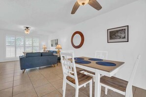 A modern living room with a sofa, dining table, and chairs. There are windows with blinds, a ceiling fan, and a framed picture on the wall. - Warm & Quaint Retreat with Pool (Tucson)