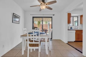 Un coin repas avec une table et des chaises, un ventilateur de plafond, une porte-fenêtre coulissante donnant sur une terrasse extérieure, et une cuisine avec des meubles en bois et un lave-vaisselle.