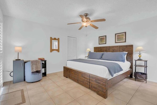 Une chambre à coucher comprenant un lit, un ventilateur de plafond, une table de chevet, une lampe, un petit meuble et une porte.