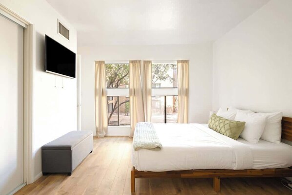 A bedroom with a bed, a bench, a TV, and a view through a window. - Midtown Retreat-Pet Friendly (Tucson)
