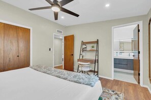 Ein Schlafzimmer mit einem Bett, einem Deckenventilator, einem Schreibtisch, einem Stuhl und einem durch eine offene Tür einsehbaren Badezimmer.
